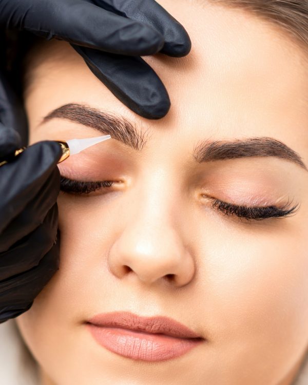 Beautician applying permanent makeup on eyebrows of young woman by special tattoo machine tool